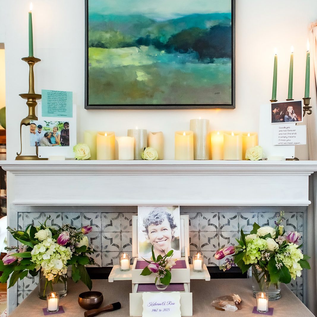 Memorial shrine on a white mantel with lit candles, fresh flowers, and photos surrounding a central portrait of a smiling woman/elderly person.